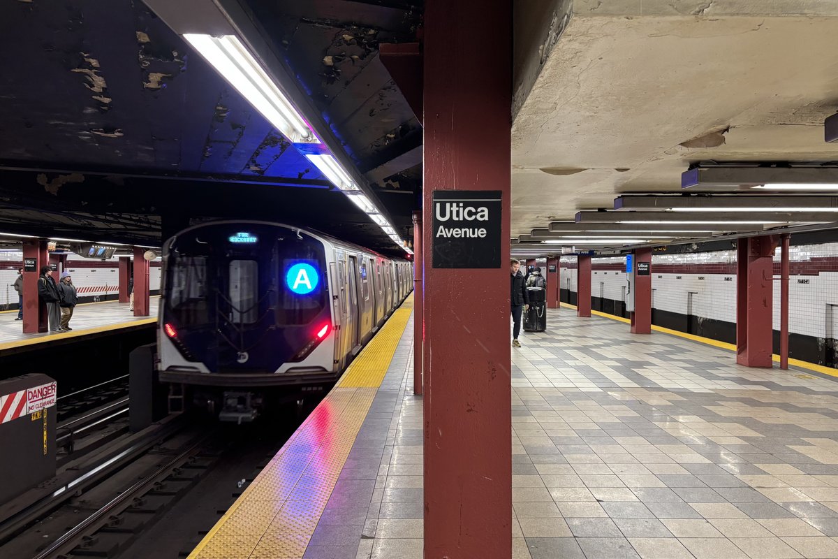 A New York City subway A train with its blue A sign lit pulling along the yellow-edged platform at Utica Avenue station in Brooklyn, beside a maroon pillar carrying the station name sign