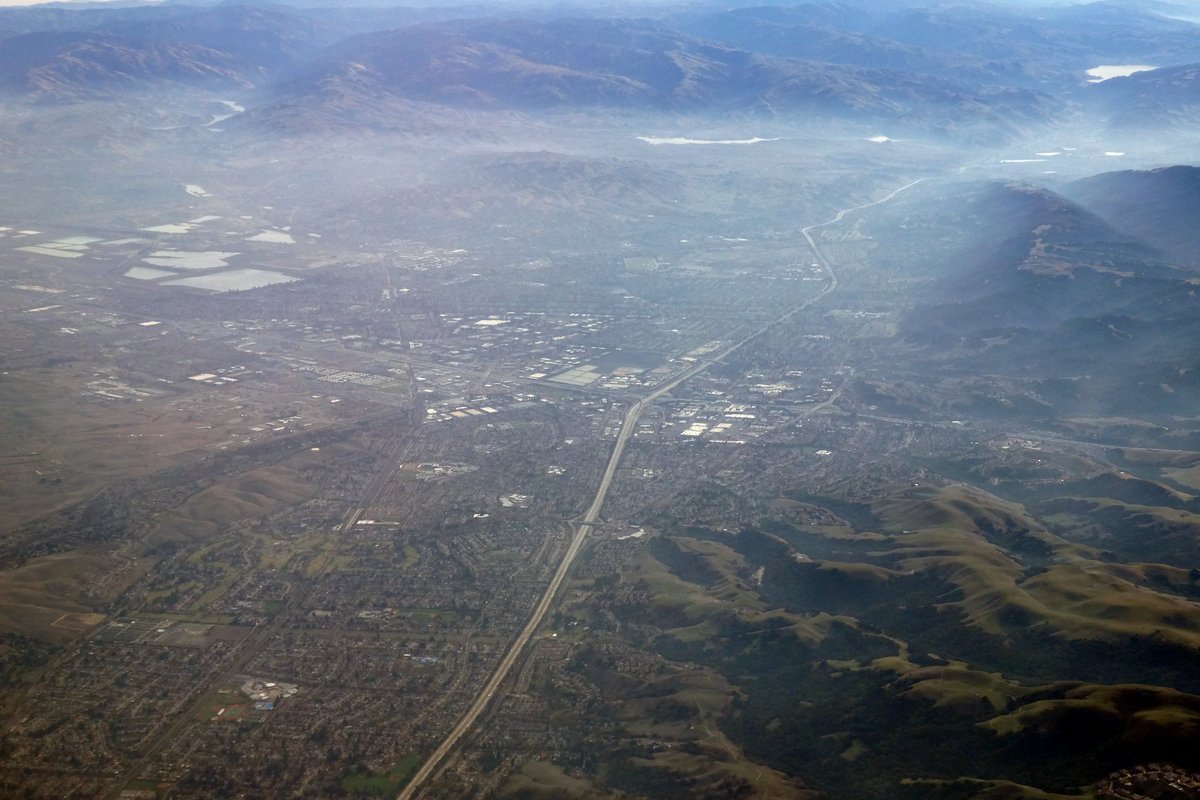 Aerial view of Dublin and Pleasanton in California's Tri-Valley, with a freeway running through suburban streets and business parks toward hazy brown hills under morning fog