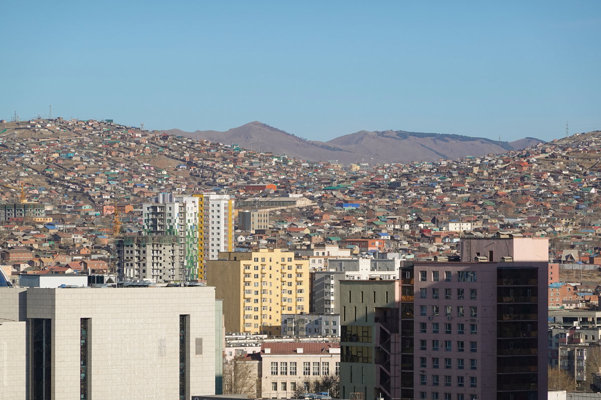 Ger districts of small houses and felt tents spreading across bare brown hills on the edge of Ulaanbaatar, Mongolia, with apartment blocks in the foreground and mountains behind under a clear blue sky