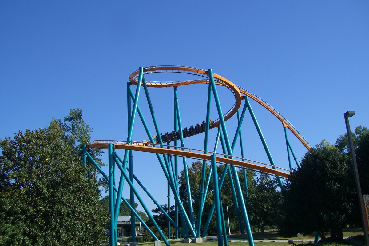 The Goliath roller coaster at Six Flags Over Georgia: teal steel supports holding up orange track that curves through a high turn against a clear blue sky, with a train of riders passing through the structure and trees on either side