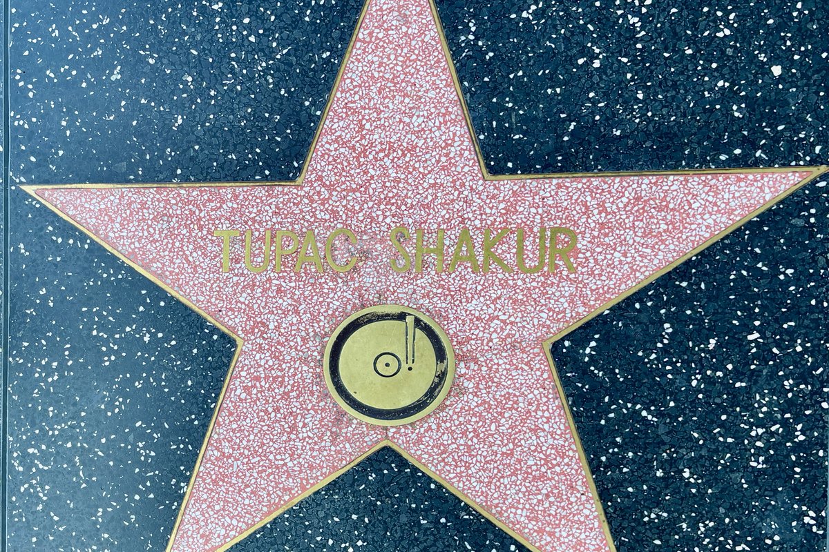 Tupac Shakur&rsquo;s star on the Hollywood Walk of Fame: a coral-pink terrazzo star edged in brass, with TUPAC SHAKUR in brass capitals above a round brass phonograph-record emblem, set into dark speckled terrazzo pavement