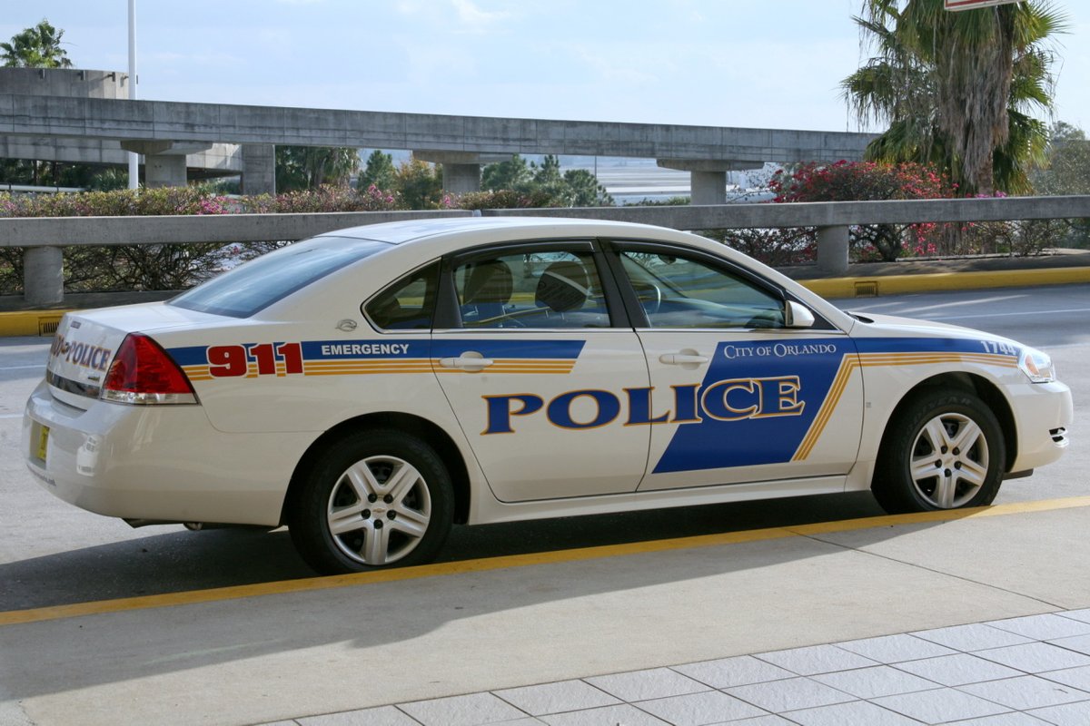 A white City of Orlando police patrol car parked at a kerb, with a blue POLICE stripe on the doors, a red 911 Emergency panel on the rear wing, palm trees and pink flowering shrubs behind a concrete overpass