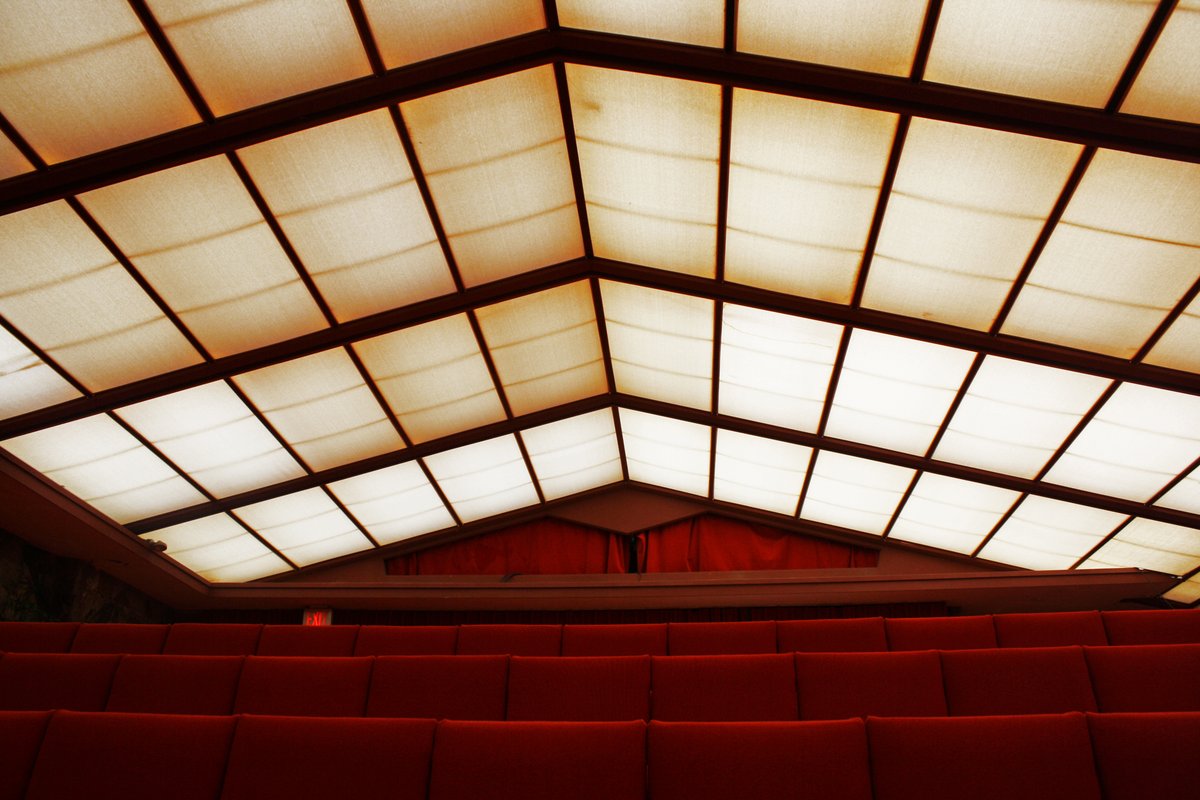 Looking up at the glowing translucent roof panels of the theatre at Taliesin West, set in a pitched grid of dark beams above rows of red theatre seats and a red stage curtain