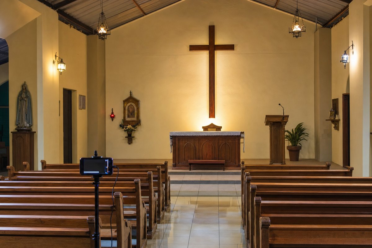 An empty parish church at dusk, with a large plain wooden cross mounted on the wall above a wooden altar, rows of wooden pews and a phone on a tripod set up near the front as if to livestream the service